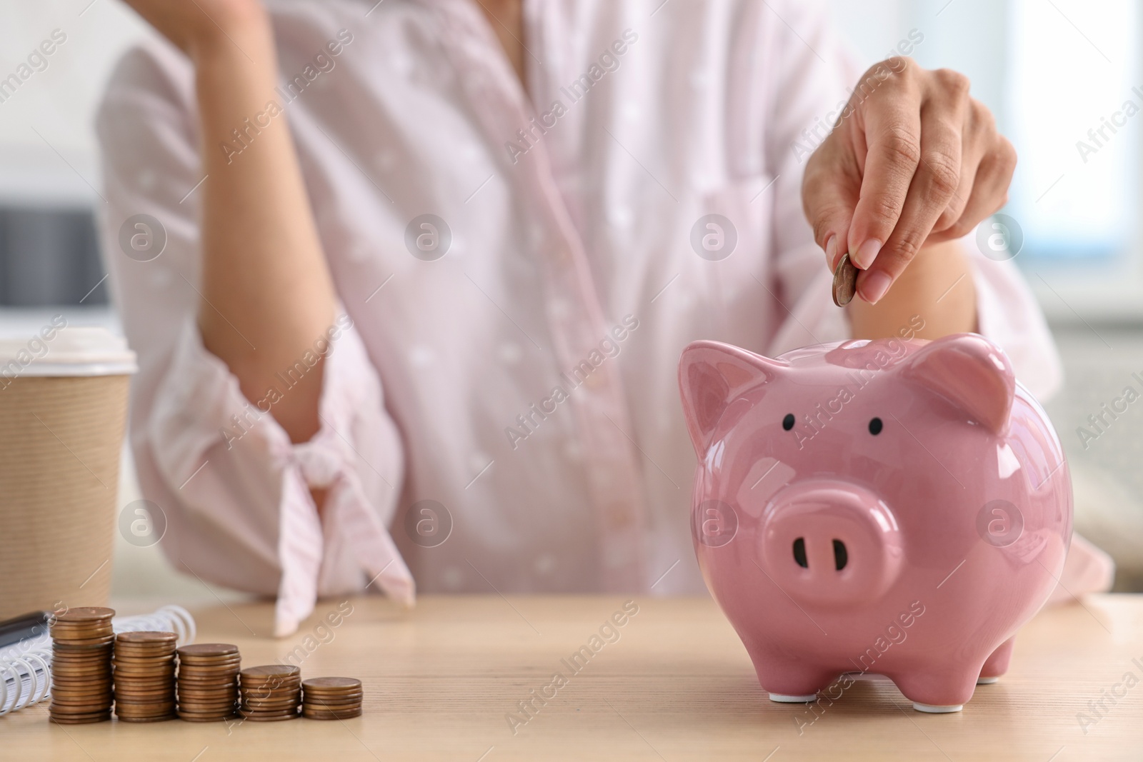 Photo of Woman putting coin into piggy bank at table indoors, closeup