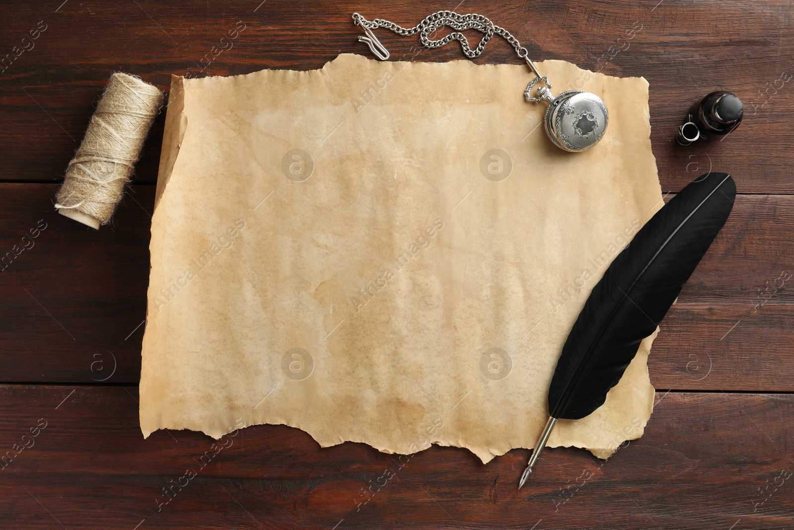 Photo of Sheet of old parchment paper, black feather, inkwell, rope and pocket chain clock on wooden table, flat lay