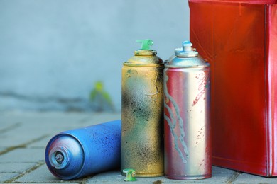 Many spray paint cans and red cardboard box outdoors, closeup. Space for text