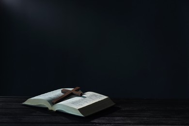 Photo of Bible and cross on black wooden table against dark background, space for text. Religion of Christianity