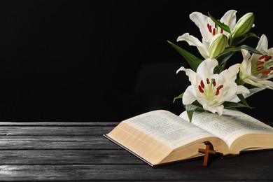 Bible, cross and lilies on dark gray wooden table against black background, space for text. Religion of Christianity