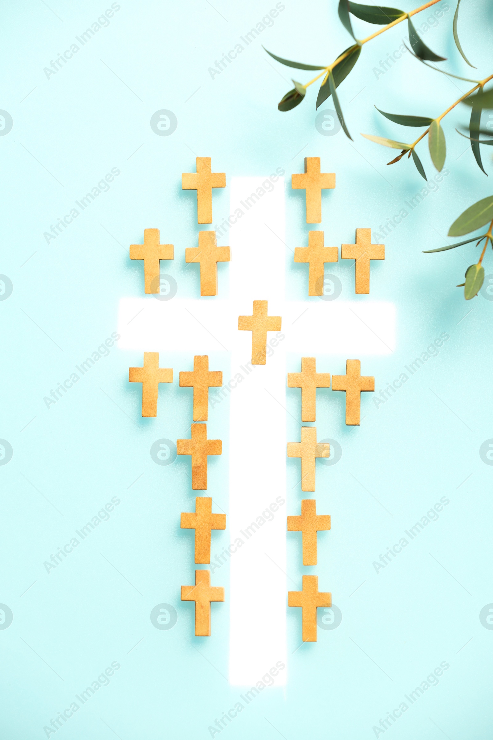 Photo of White cross among small ones and eucalyptus branches on turquoise background, flat lay. Religion of Christianity