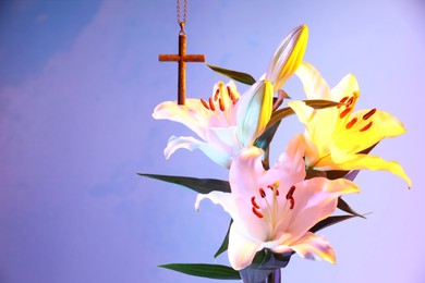 Photo of One cross with chain and beautiful lily flowers on violet background, space for text. Religion of Christianity