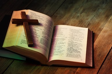 Photo of Cross and Bible on wooden table in color lights, closeup. Religion of Christianity