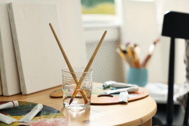 Artist's palette, brushes in glass of water and paints on wooden table indoors