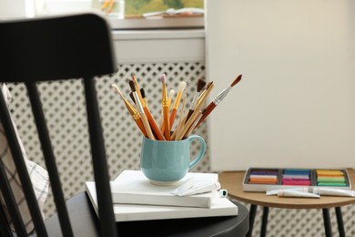 Photo of Canvases, paints and cup of brushes on chair in workshop