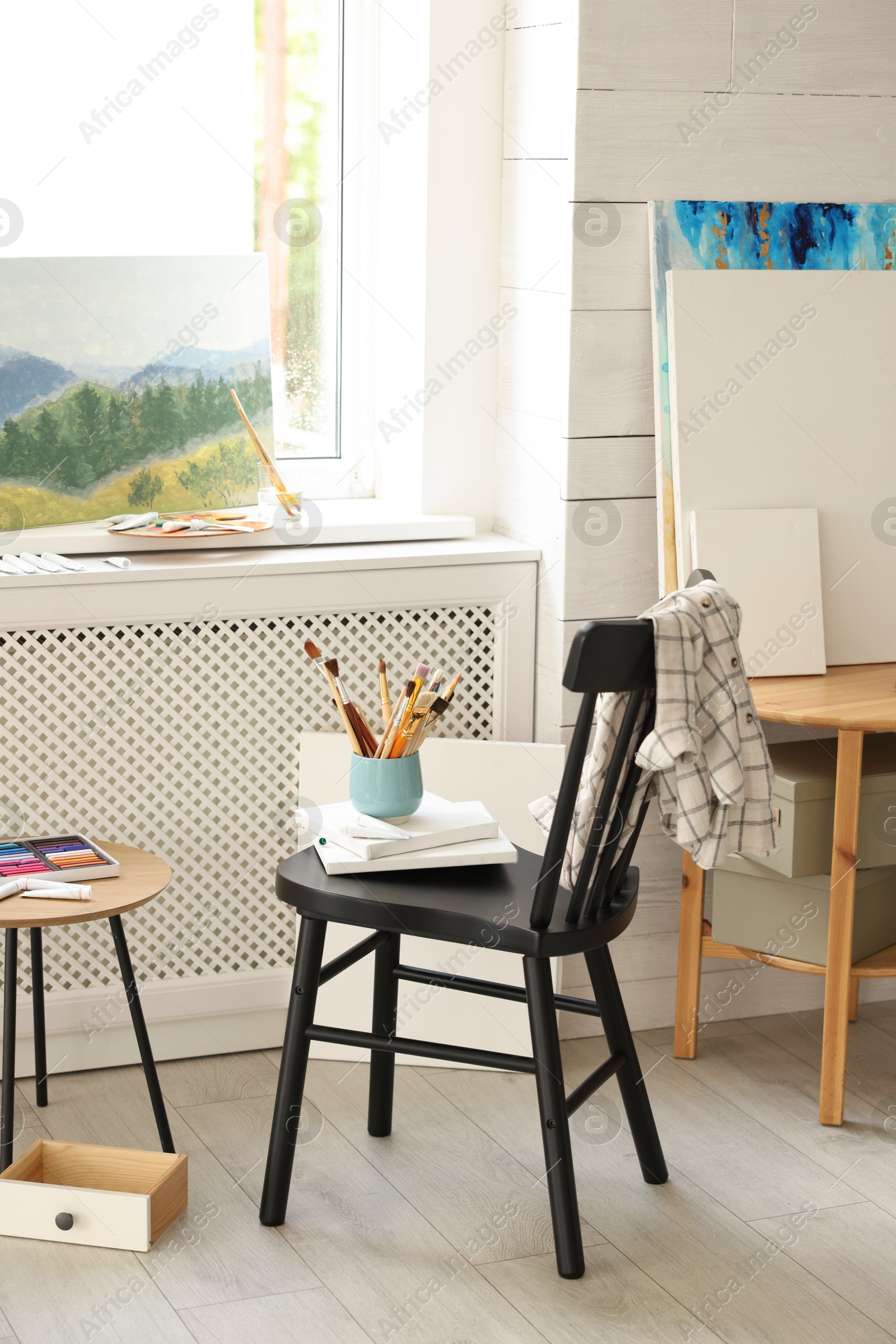 Photo of Canvases, paints and cup of brushes on chair in workshop