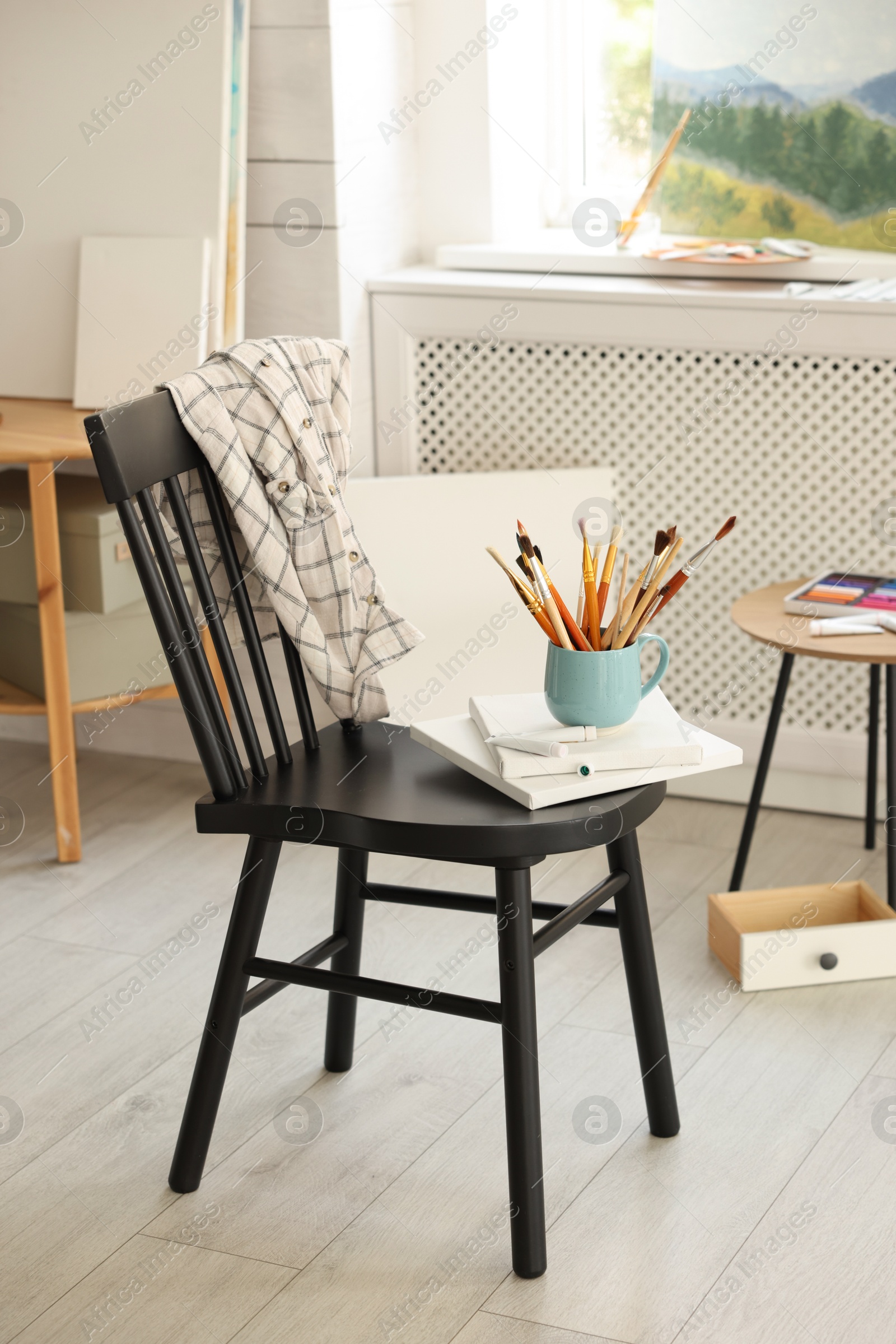 Photo of Canvases, paints and cup of brushes on chair in workshop