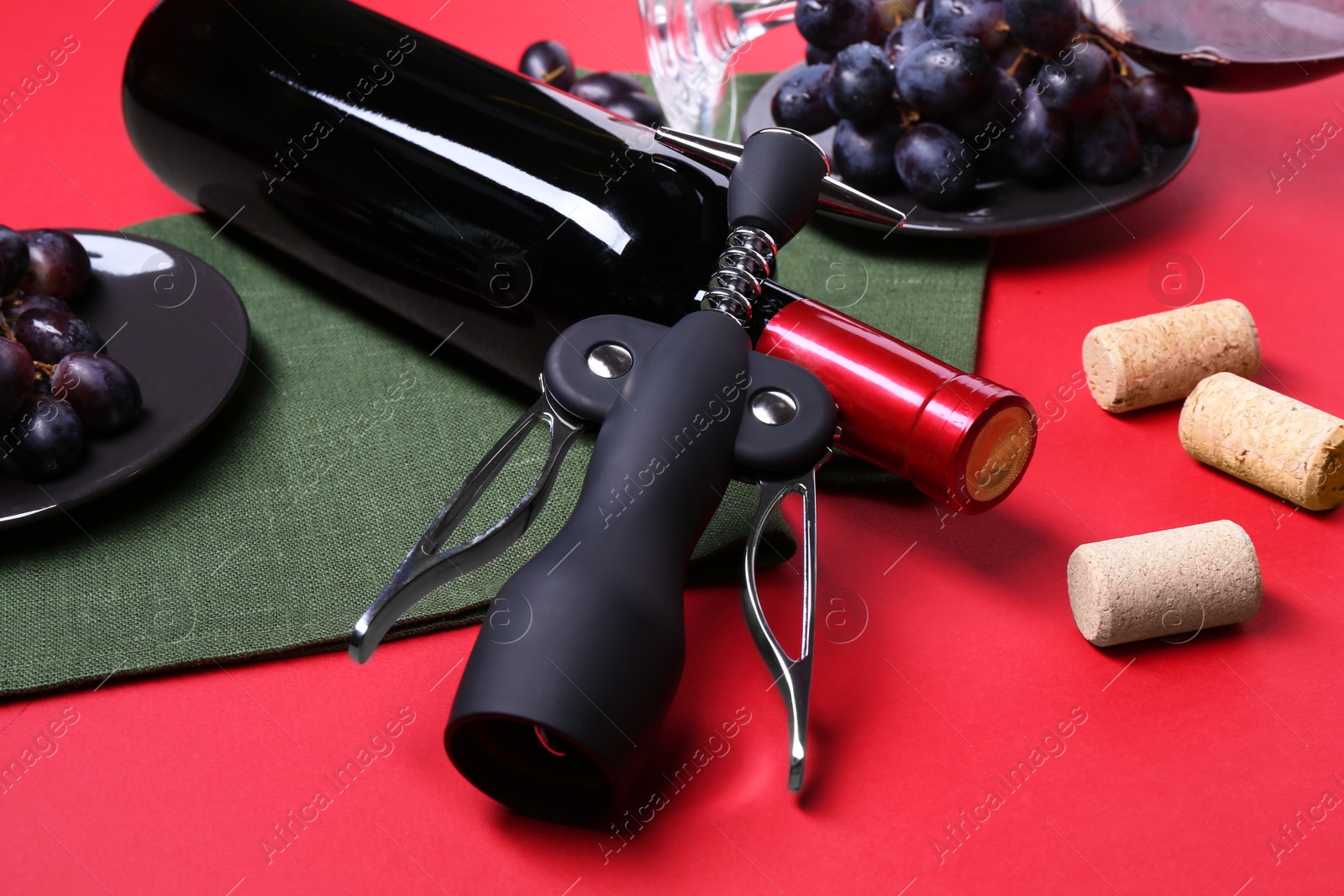 Photo of Wing corkscrew, bottle of wine, grapes and corks on red background, closeup