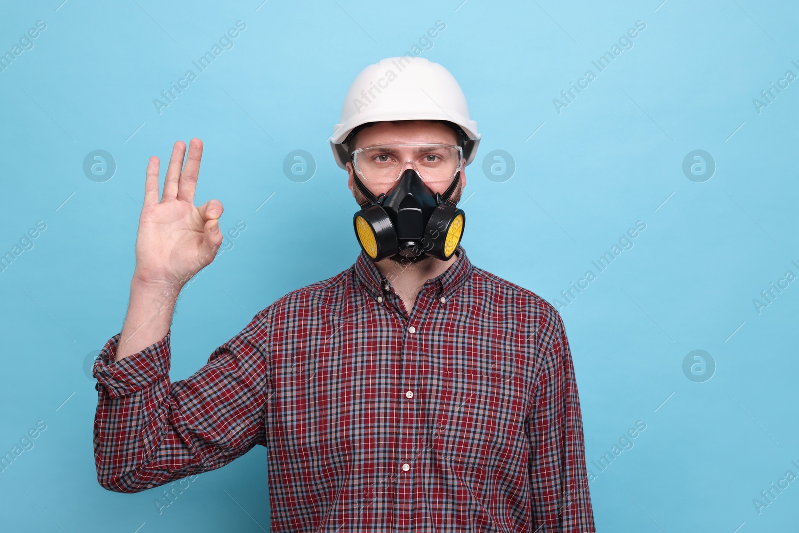 Photo of Man in respirator mask and hard hat showing OK gesture on light blue background