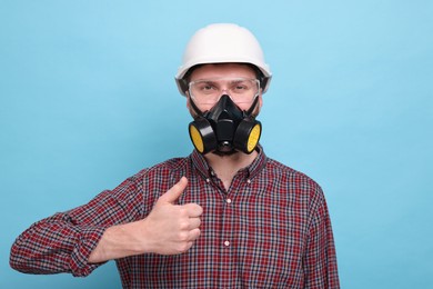 Photo of Man in respirator mask and hard hat showing thumb up on light blue background