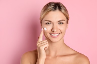 Beautiful woman applying cream on her face against pink background