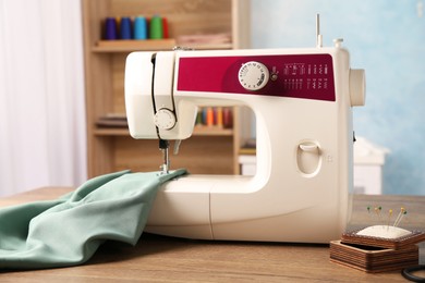 Photo of White sewing machine, color fabric and pin cushion with needles on wooden table indoors