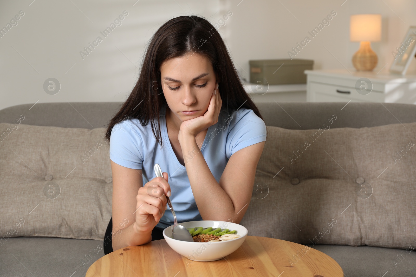 Photo of Eating disorder. Sad woman holding spoon near bowl with granola, kiwi and banana at wooden table indoors