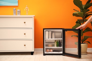 Photo of Woman opening mini refrigerator with drinks and snacks indoors, closeup