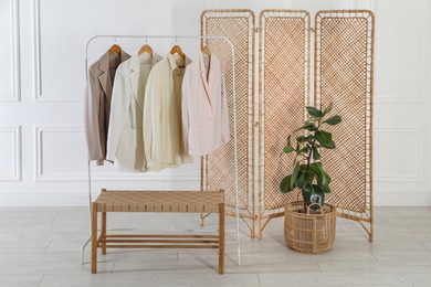 Photo of Folding screen, rack with clothes, storage bench and houseplant near white wall indoors