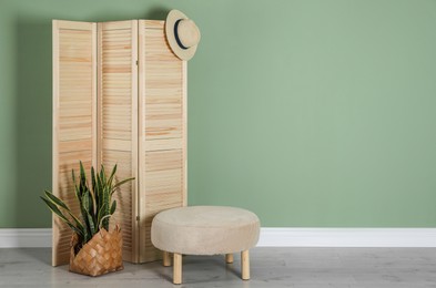 Photo of Wooden folding screen, pouffe, bag, houseplant and hat near green wall indoors, space for text