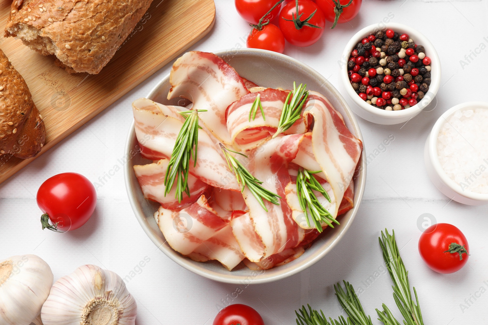 Photo of Slices of raw bacon, spices, bread and tomatoes on white tiled table, flat lay