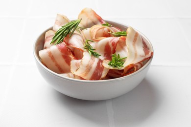 Photo of Slices of raw bacon and rosemary in bowl on white tiled table