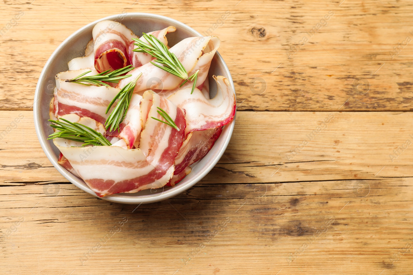 Photo of Slices of raw bacon and rosemary in bowl on wooden table, top view. Space for text