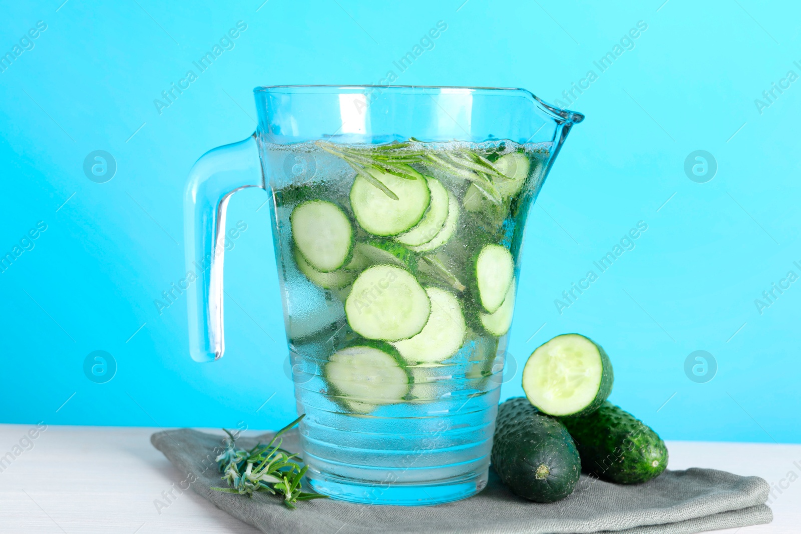 Photo of Refreshing cucumber water with rosemary in jug and vegetables on white table against light blue background, closeup