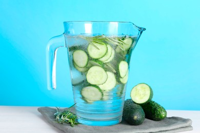 Photo of Refreshing cucumber water with rosemary in jug and vegetables on white table against light blue background, closeup