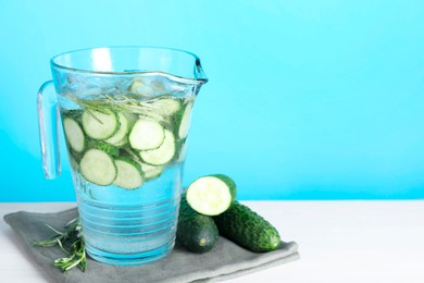 Photo of Refreshing cucumber water with rosemary in jug and vegetables on white table against light blue background. Space for text