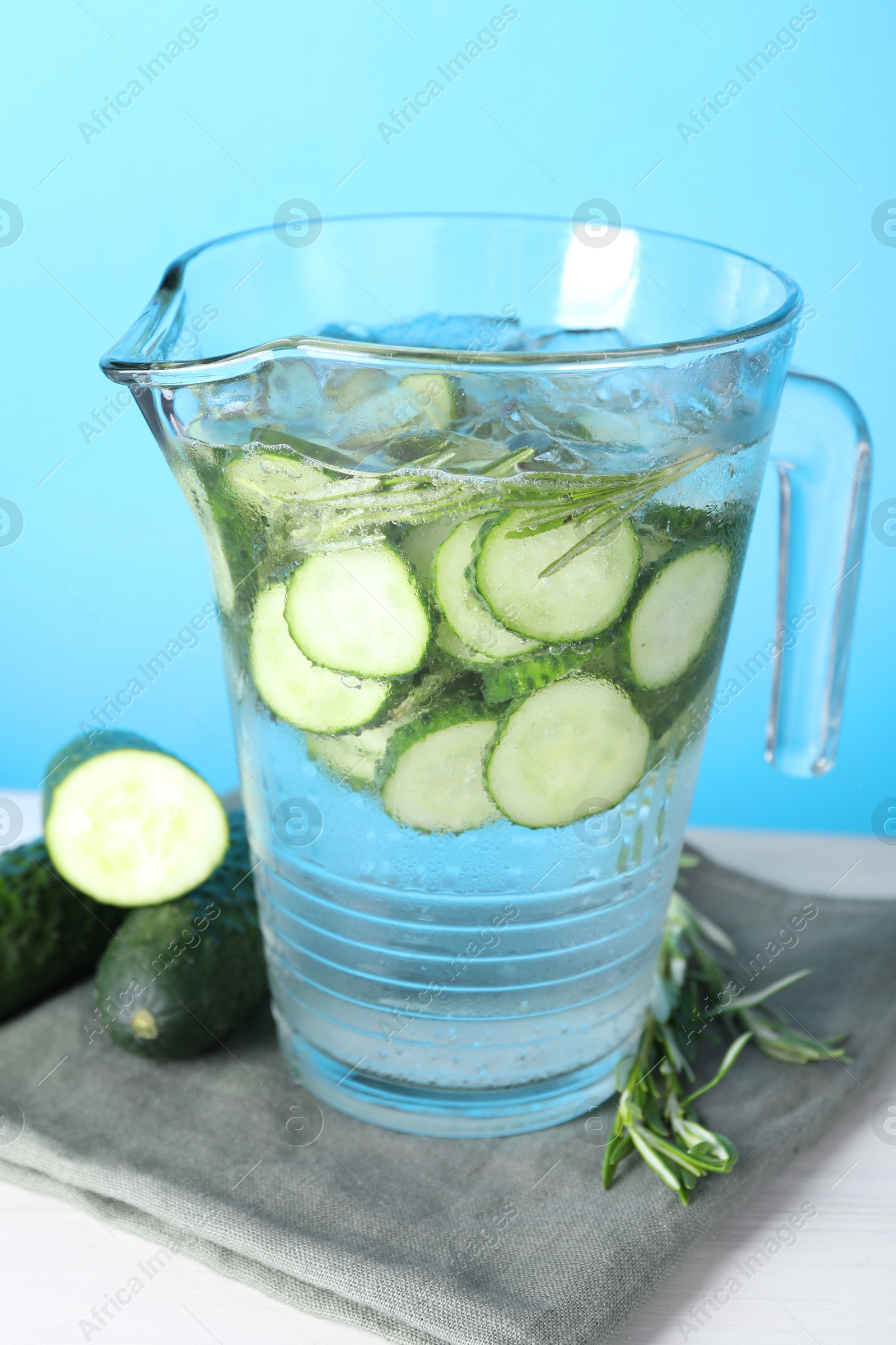 Photo of Refreshing cucumber water with rosemary in jug and vegetables on white table against light blue background