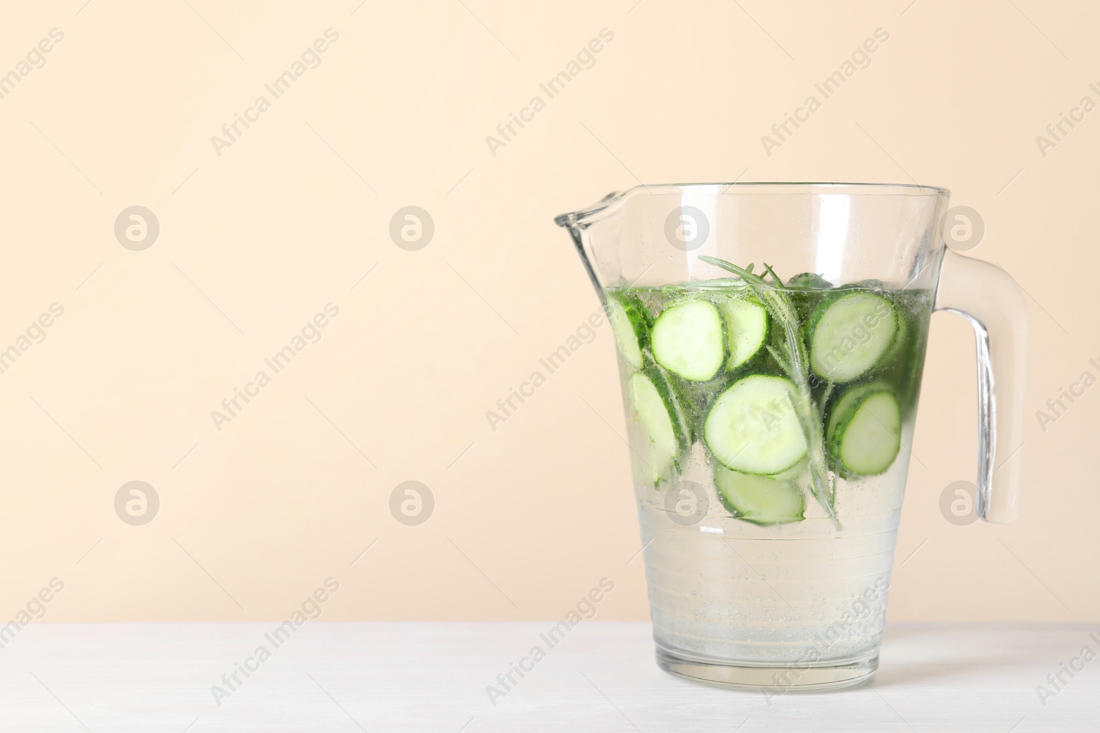 Photo of Refreshing cucumber water with rosemary in jug on white table against beige background. Space for text