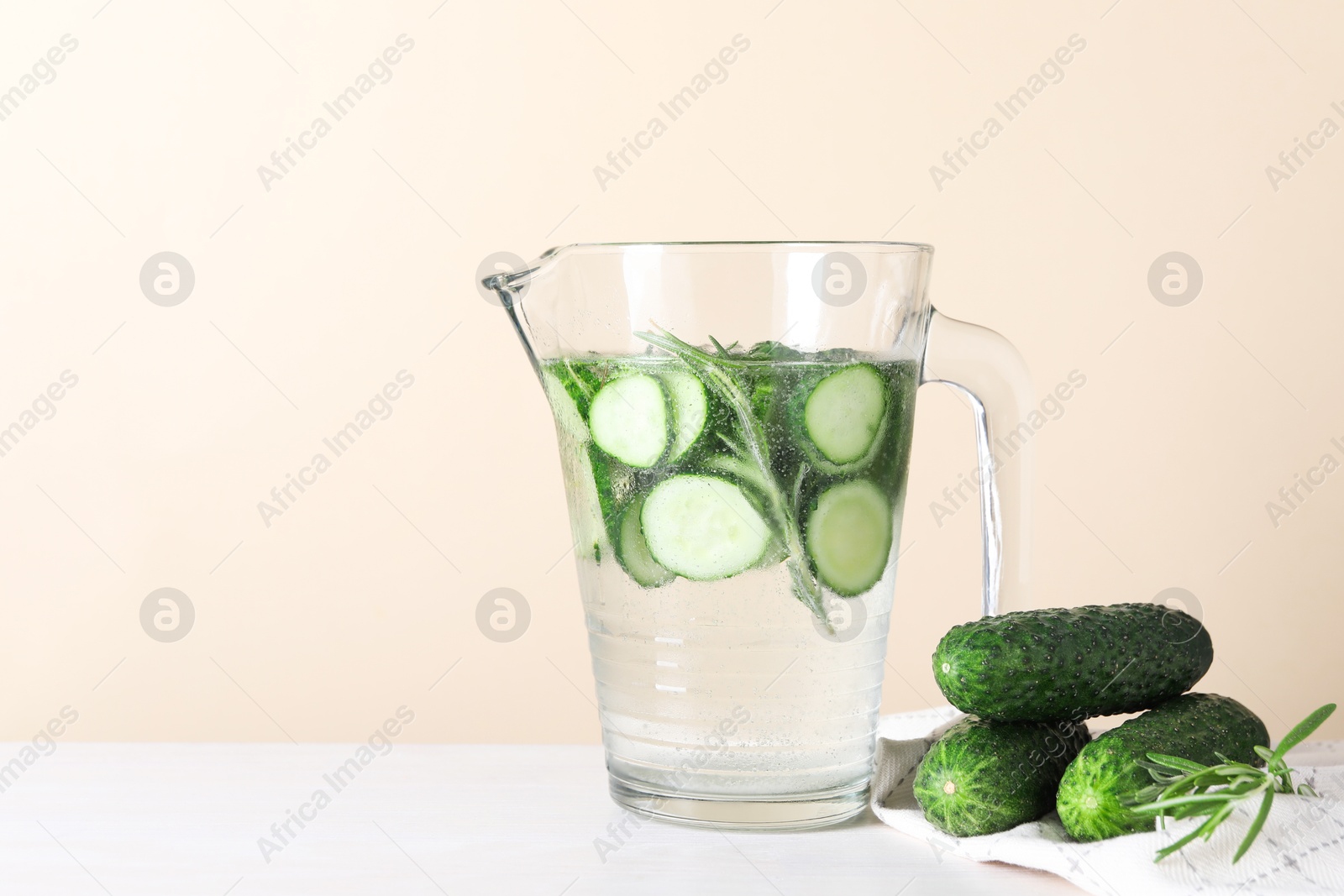 Photo of Refreshing cucumber water with rosemary in jug and vegetables on white table against beige background. Space for text