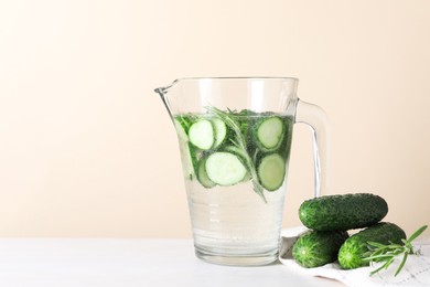 Photo of Refreshing cucumber water with rosemary in jug and vegetables on white table against beige background. Space for text