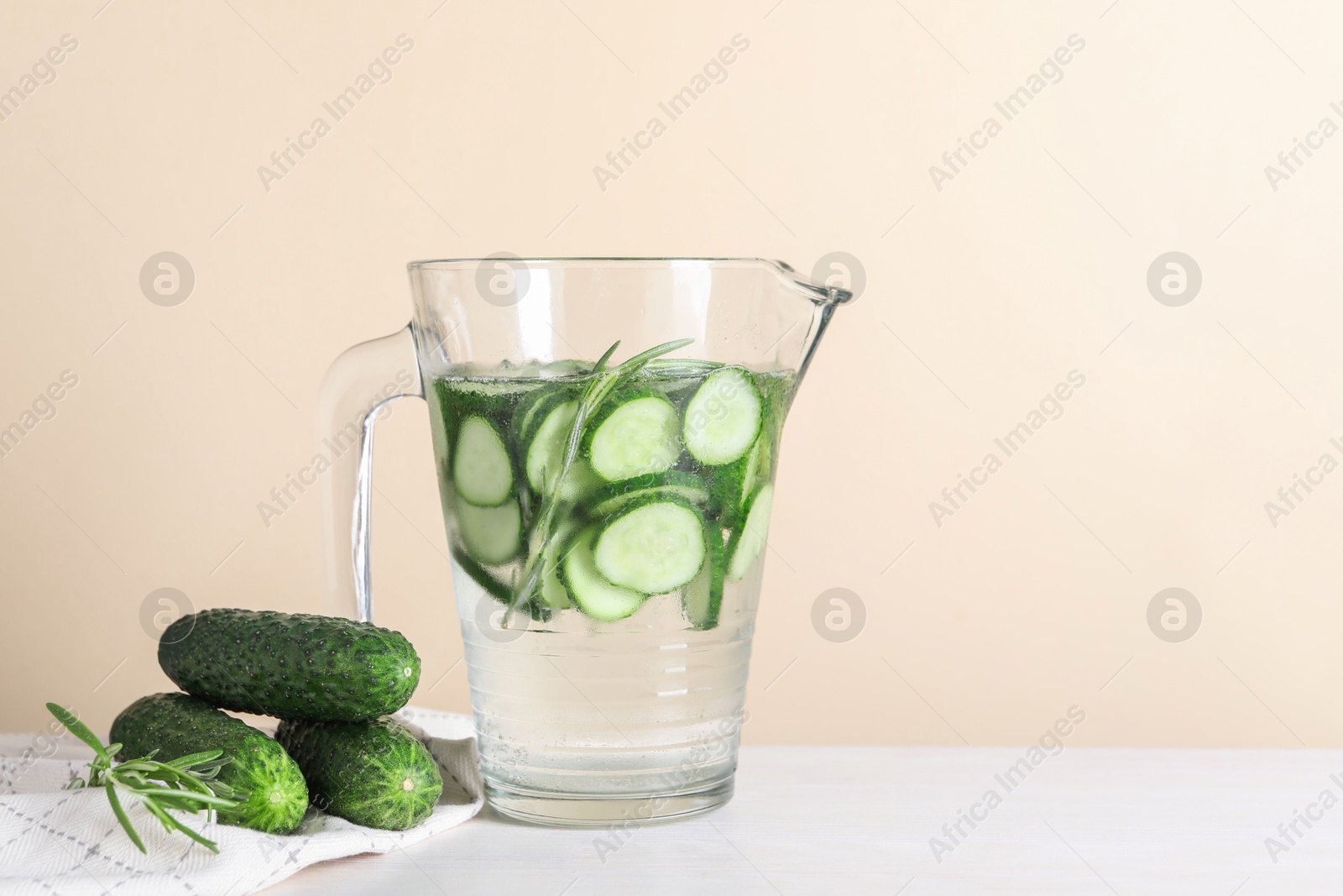 Photo of Refreshing cucumber water with rosemary in jug and vegetables on white table against beige background. Space for text