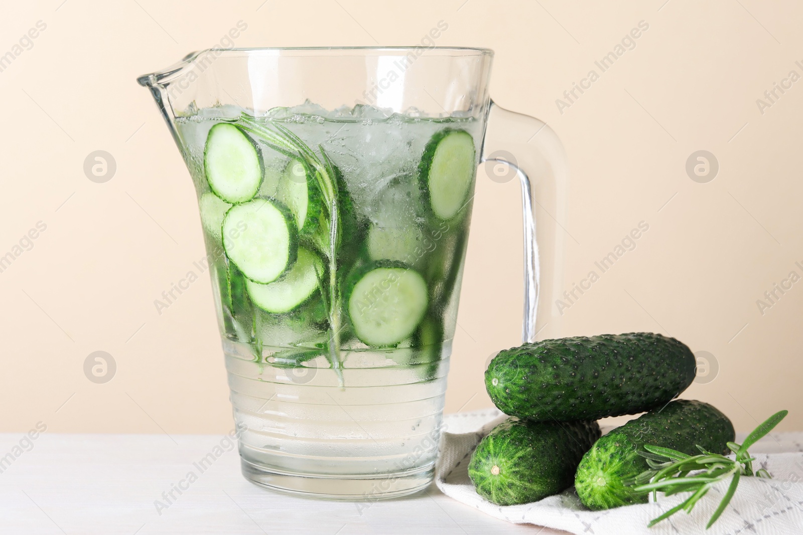 Photo of Refreshing cucumber water with rosemary in jug and vegetables on white table against beige background, closeup