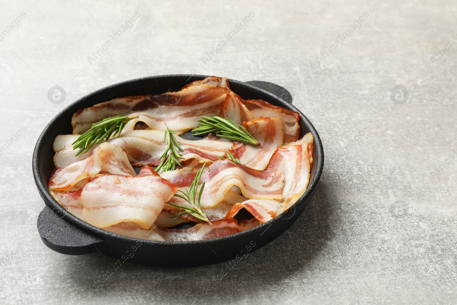 Photo of Delicious bacon slices and fresh rosemary in frying pan on light grey table. Space for text