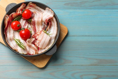 Delicious bacon slices, spices and tomatoes in frying pan on light blue wooden table, top view. Space for text