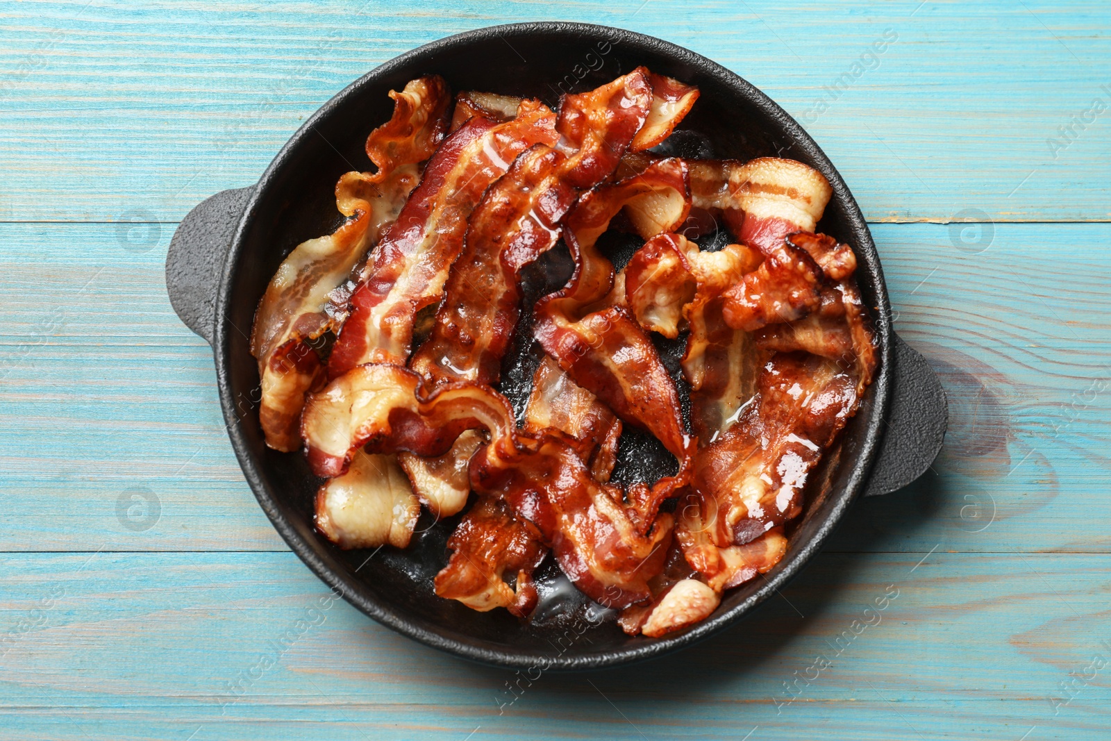 Photo of Delicious bacon slices in frying pan on light blue wooden table, top view