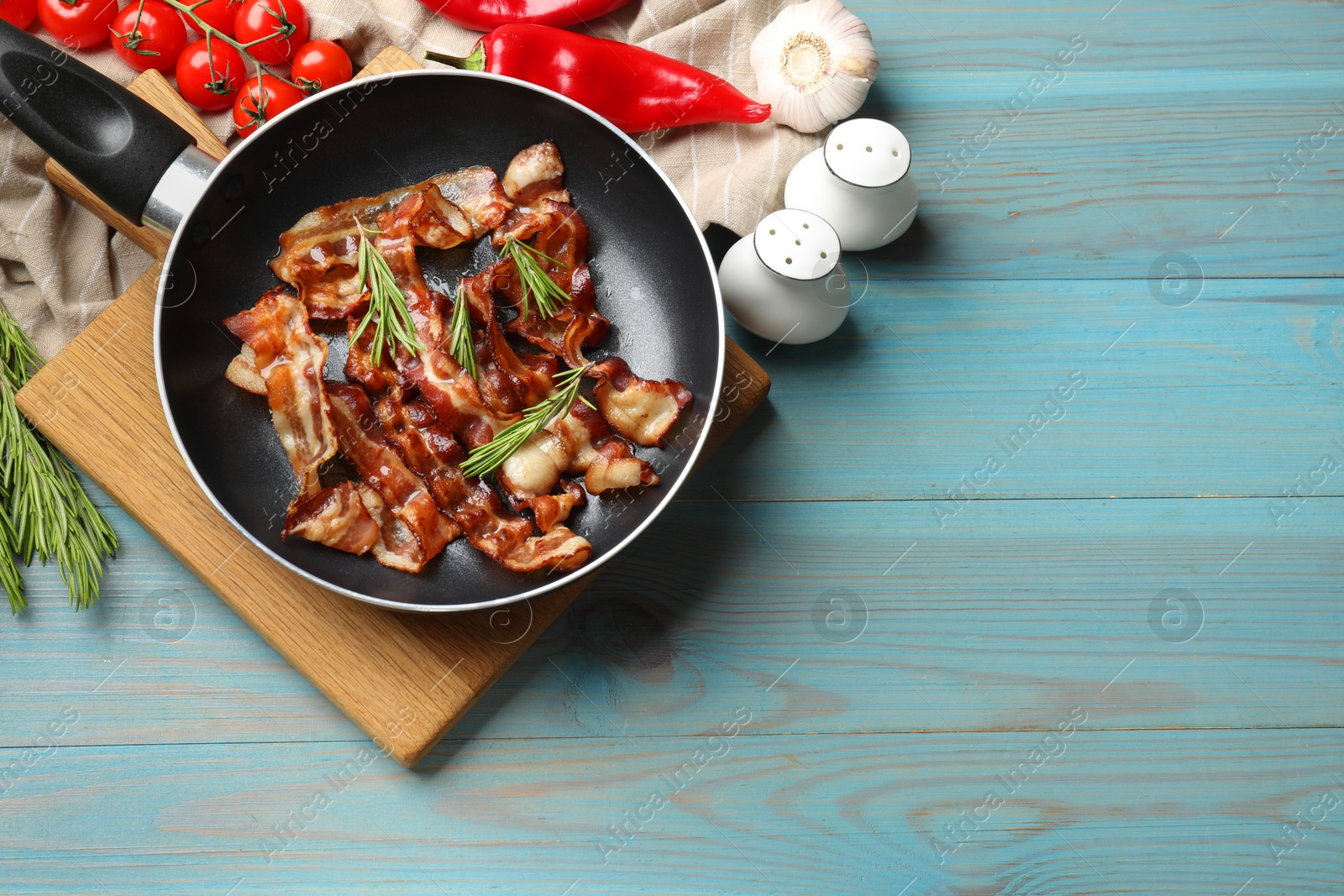 Photo of Delicious bacon slices in frying pan and products on light blue wooden table, flat lay. Space for text