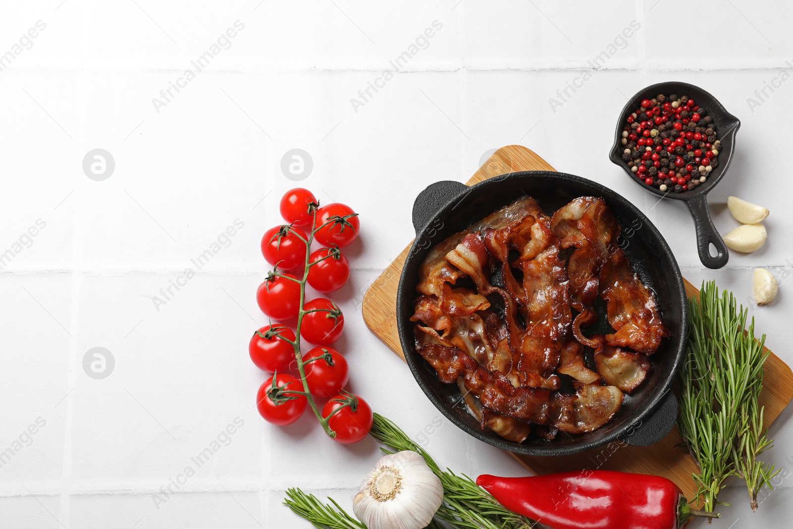 Photo of Delicious bacon slices in frying pan and products on white tiled table, flat lay. Space for text