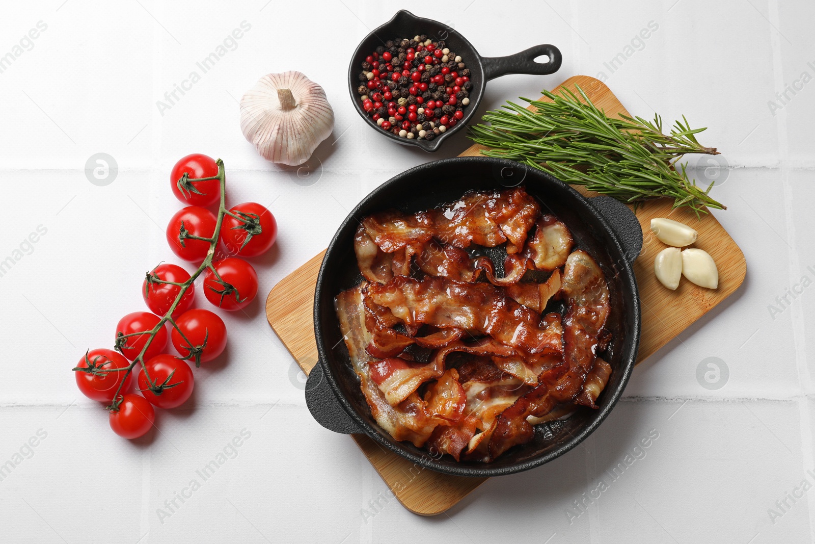 Photo of Delicious bacon slices in frying pan and products on white tiled table, flat lay