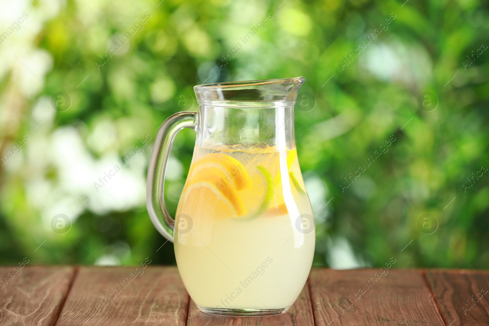 Photo of Refreshing lemonade with orange and lime in jug on wooden table against blurred green background