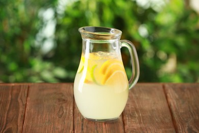 Photo of Refreshing lemonade with orange and lime in jug on wooden table against blurred green background