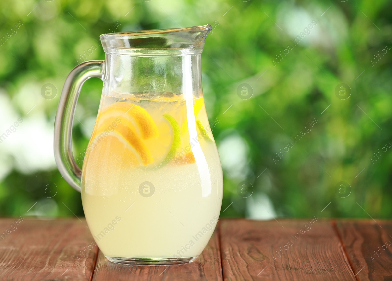 Photo of Refreshing lemonade with orange and lime in jug on wooden table against blurred green background, closeup. Space for text