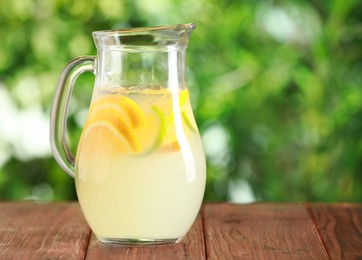 Photo of Refreshing lemonade with orange and lime in jug on wooden table against blurred green background, closeup. Space for text