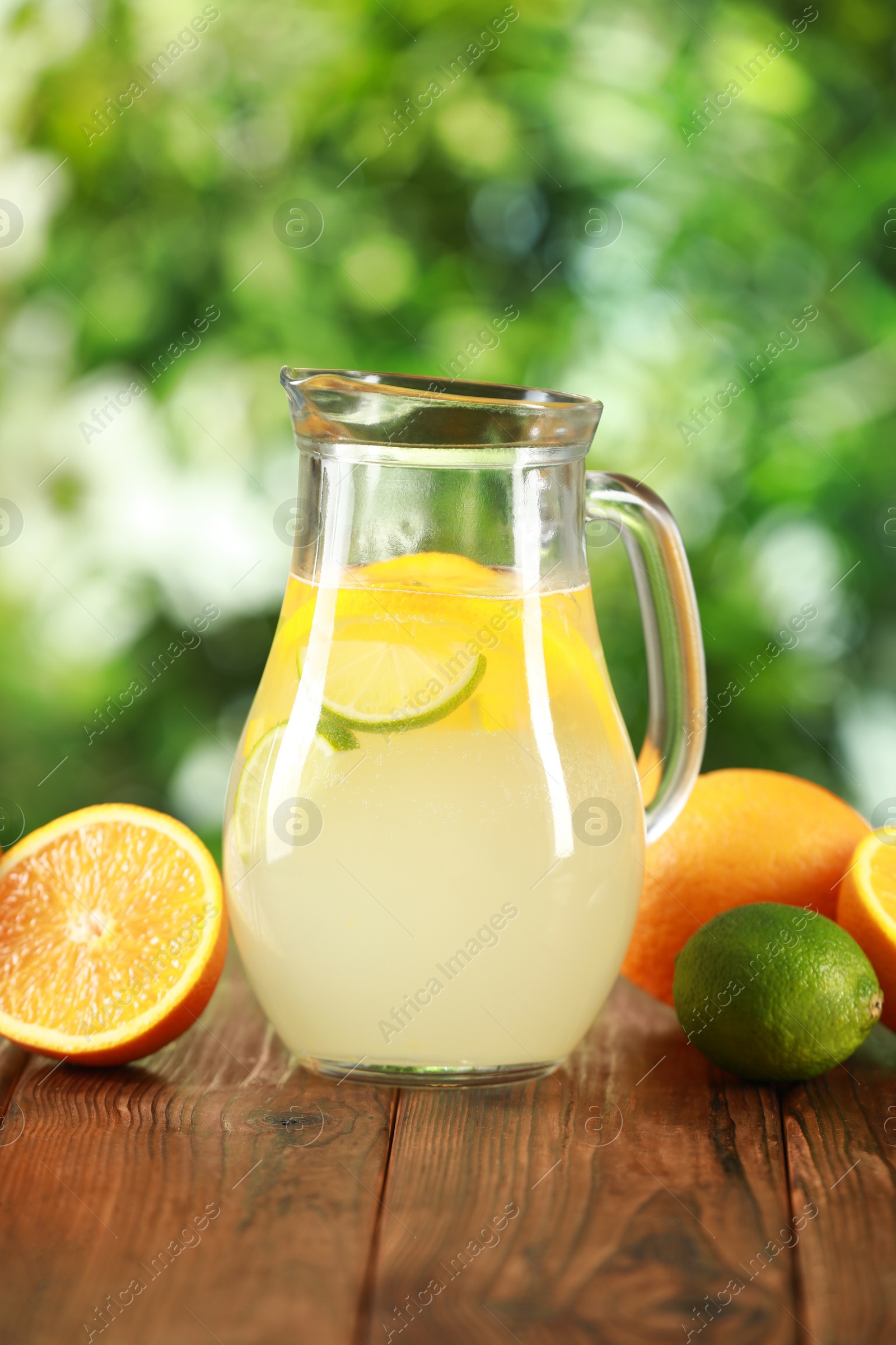 Photo of Refreshing lemonade with citruses in jug among fruits on wooden table against blurred green background