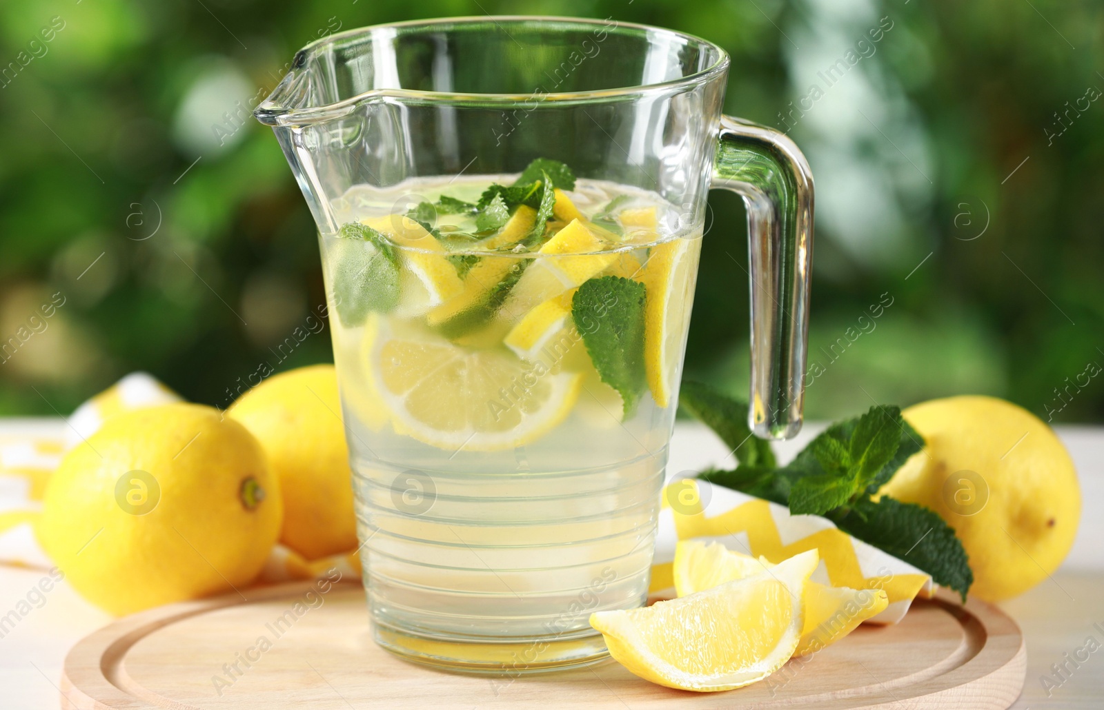 Photo of Refreshing lemonade with mint in jug among fruits on light table against blurred green background, closeup