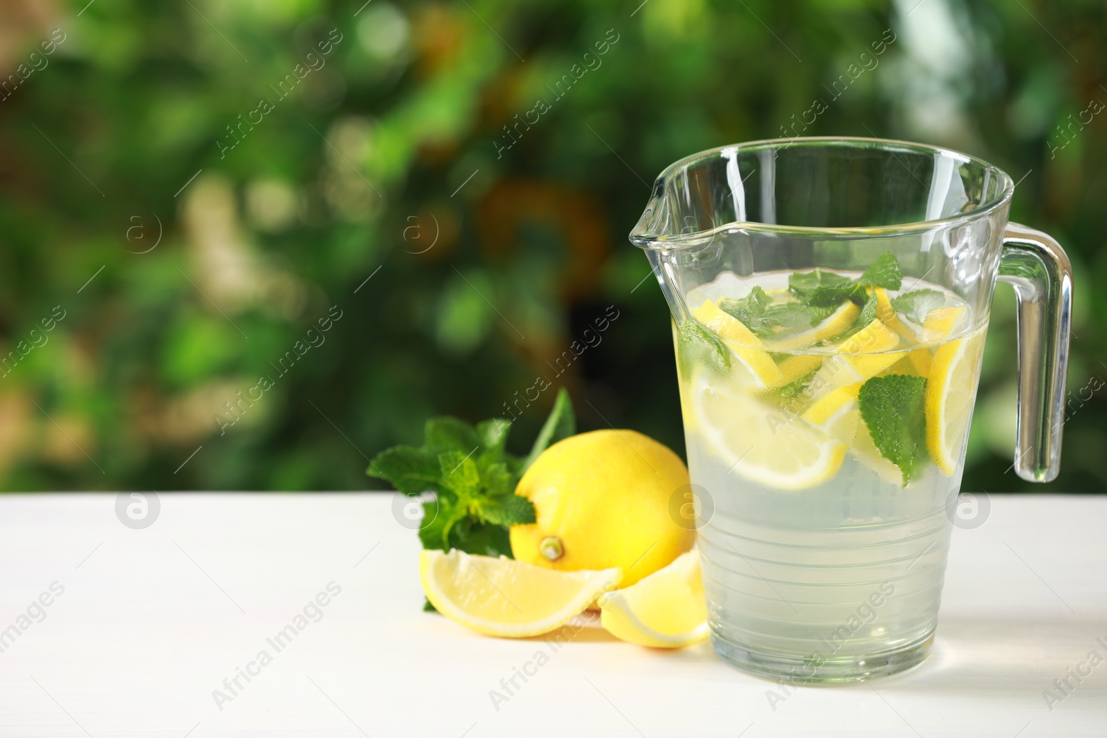 Photo of Refreshing lemonade with mint in jug on light table against blurred green background. Space for text