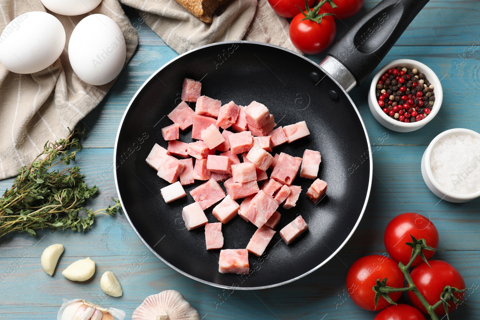 Photo of Pieces of fresh bacon in frying pan and products on light blue table, flat lay