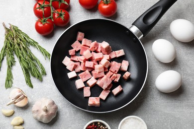 Photo of Pieces of fresh bacon in frying pan and products on grey textured table, flat lay