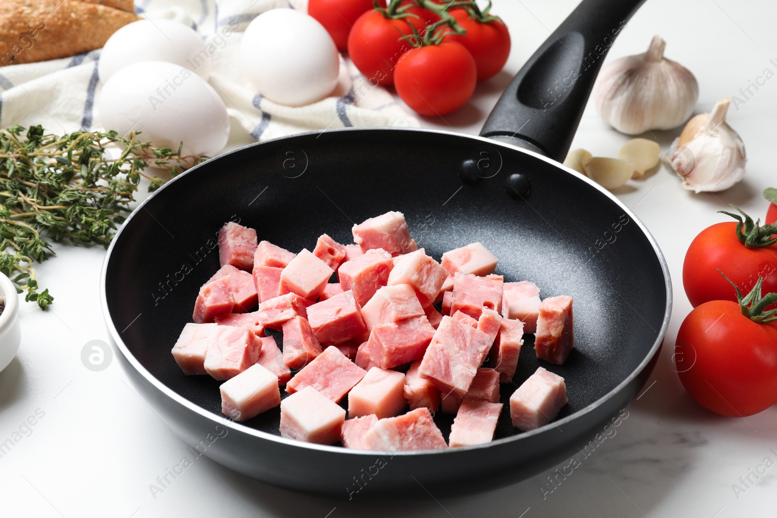 Photo of Pieces of fresh bacon in frying pan and tomatoes on white table, closeup