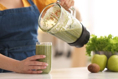 Photo of Woman pouring fresh smoothie from blender cup into glass at white marble table in kitchen, closeup